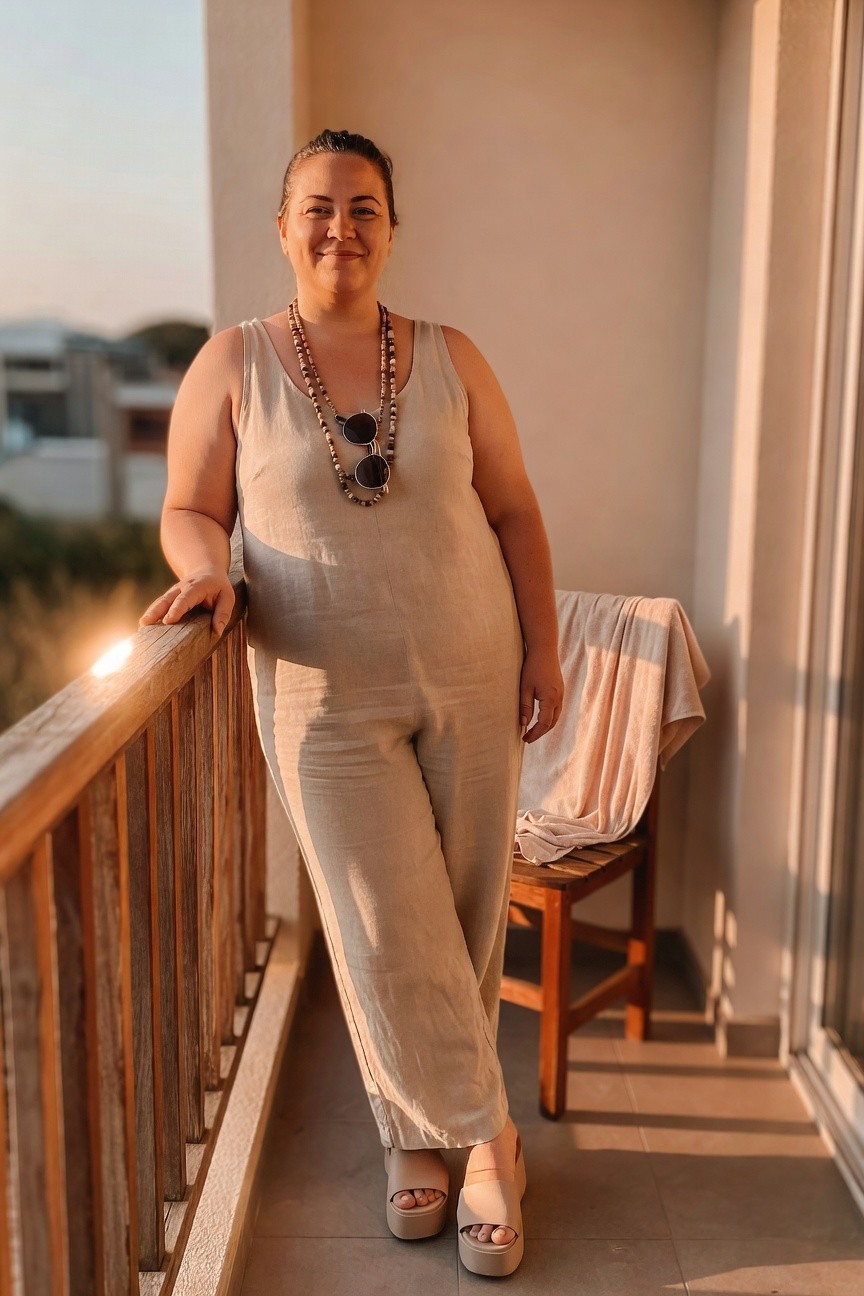 Woman in beige jumpsuit standing on balcony at sunset.