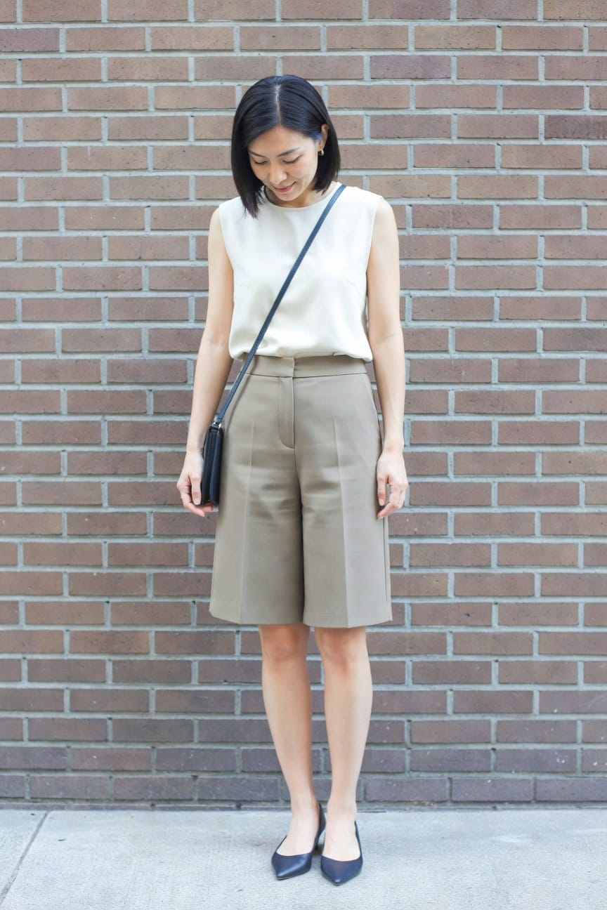Woman in cream top and taupe shorts with navy heels and crossbody bag.