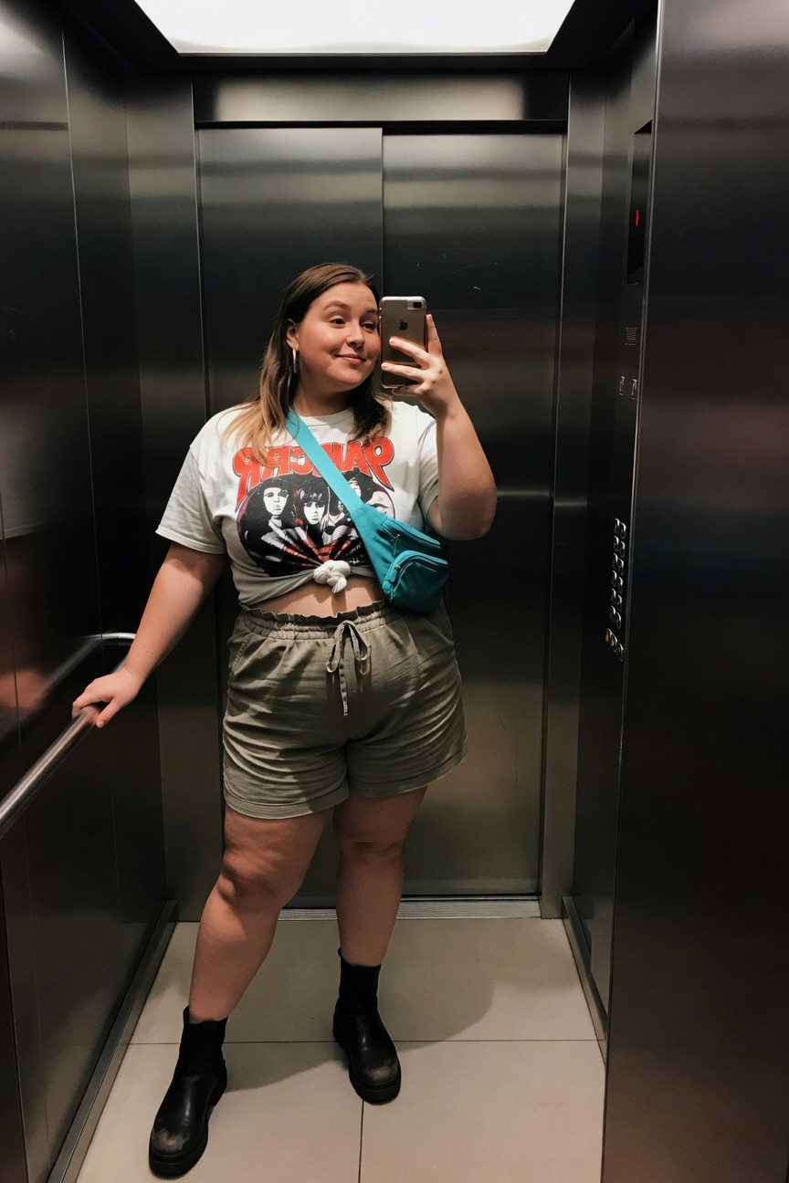 Woman in graphic tee and shorts taking mirror selfie in elevator.