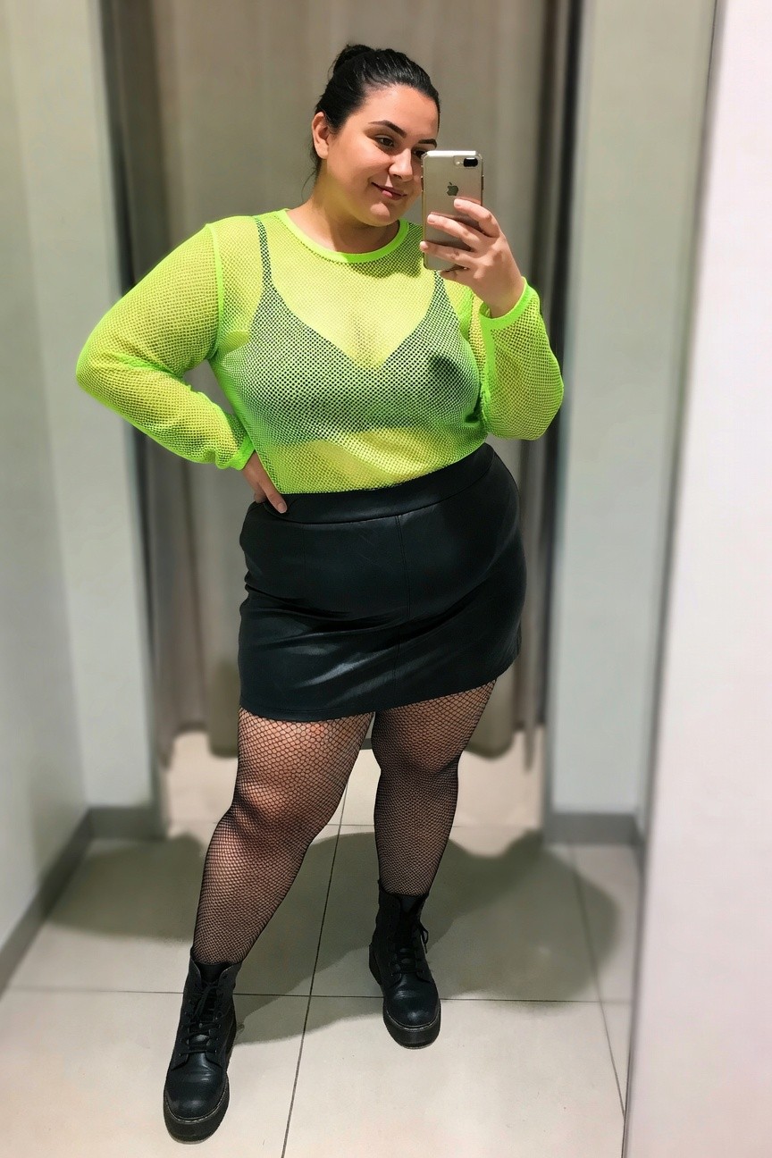 Woman in neon fishnet top and black leather skirt taking a mirror selfie.