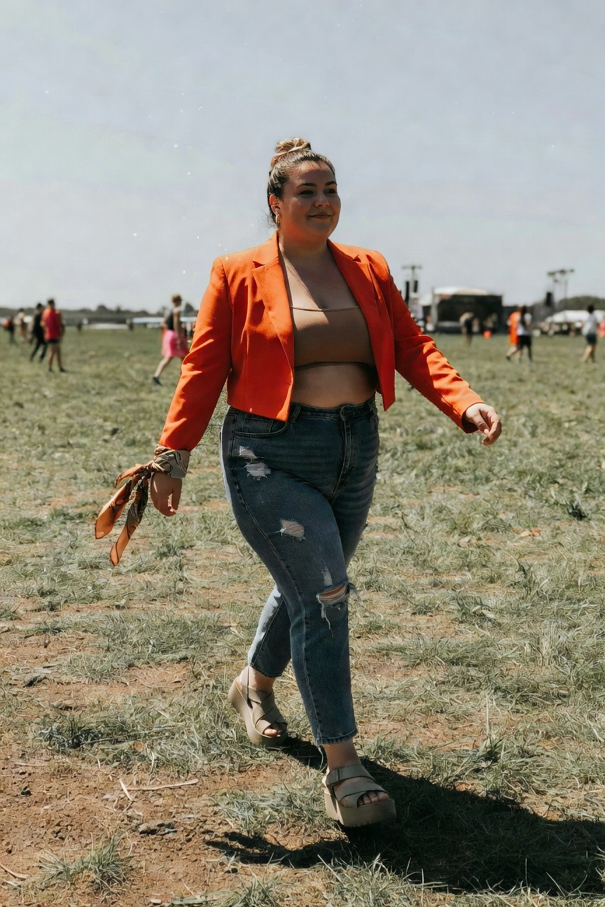 Woman in orange blazer and ripped jeans walking through sunny field.