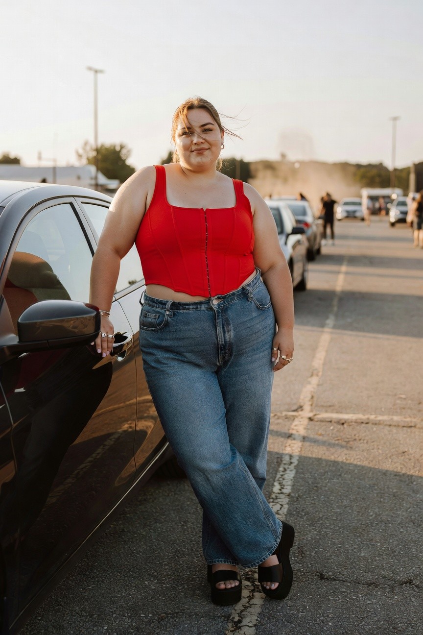 Woman in red corset top and blue jeans leaning on black car.