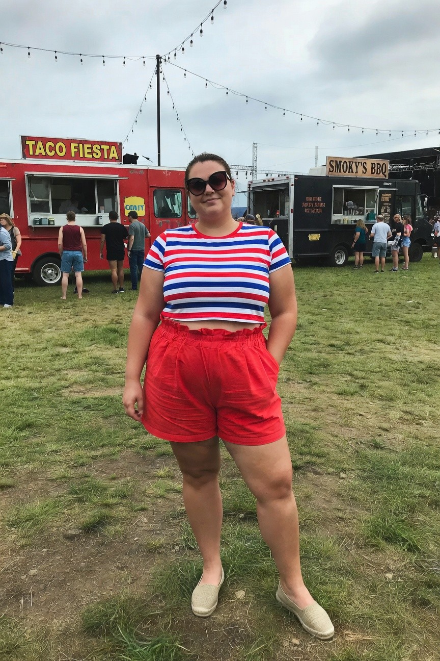 Plus size woman in a red white blue striped crop top red high waisted shorts and beige flat shoes standing in a grassy outdoor area near food trucks.