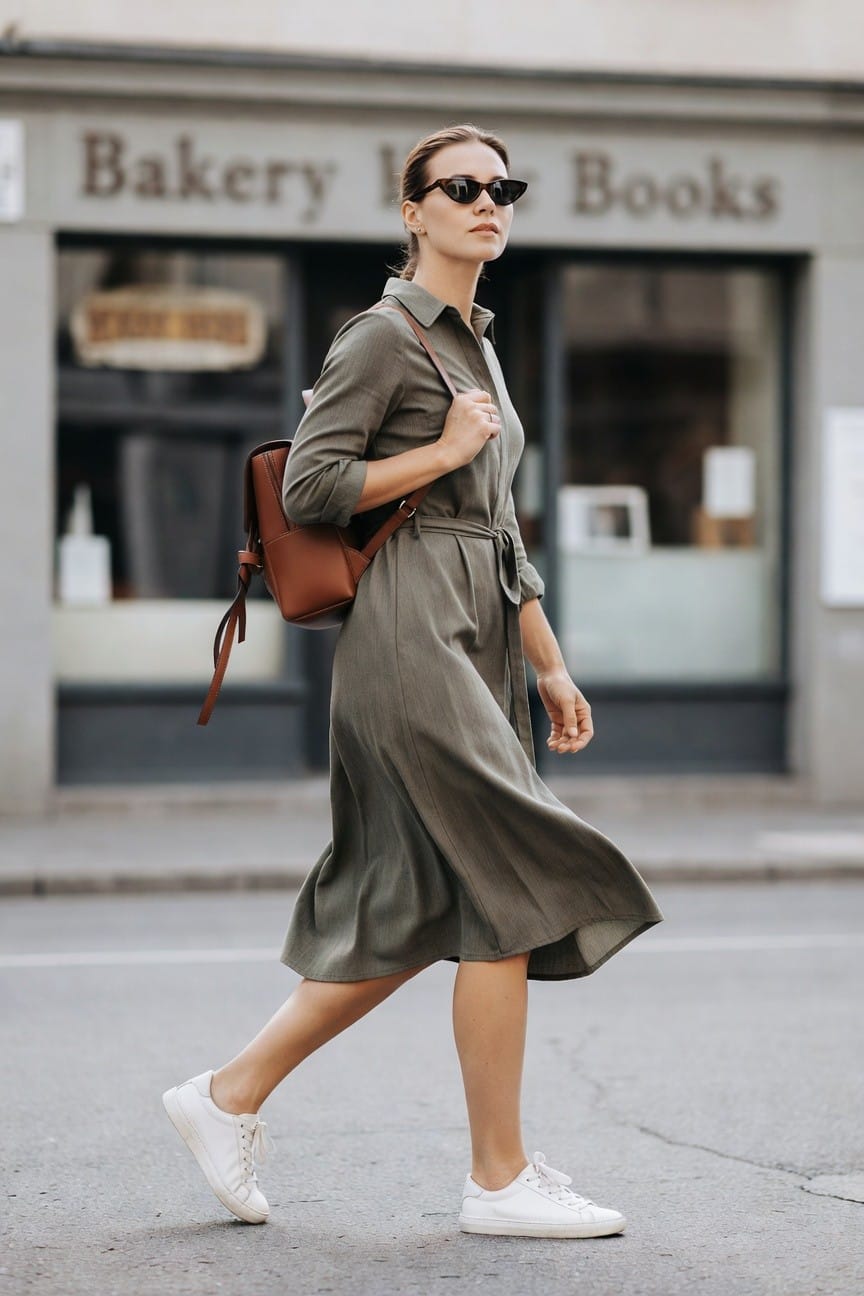 Woman in olive midi dress and white sneakers carrying brown backpack.