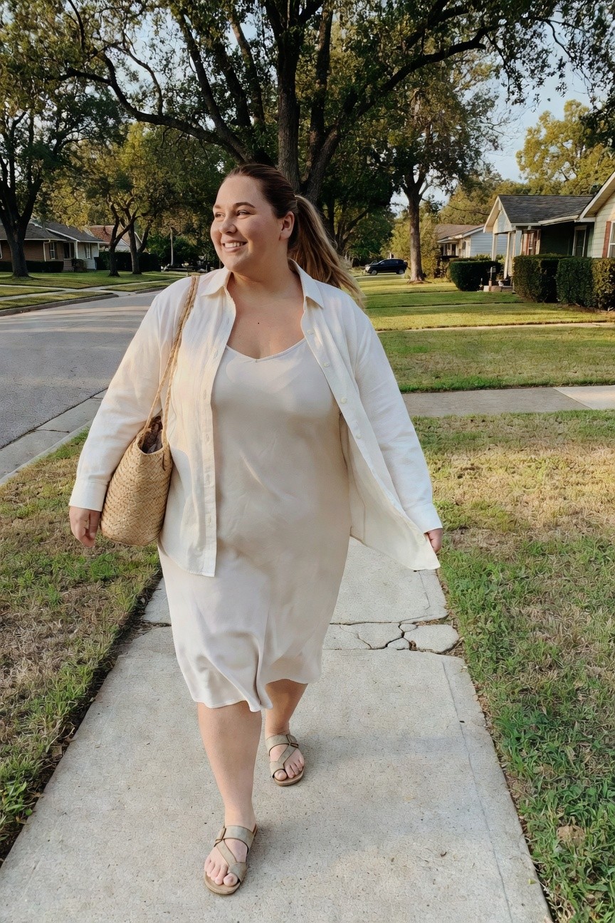 Smiling woman in cream slip dress and oversized button-up shirt walking on sidewalk.
