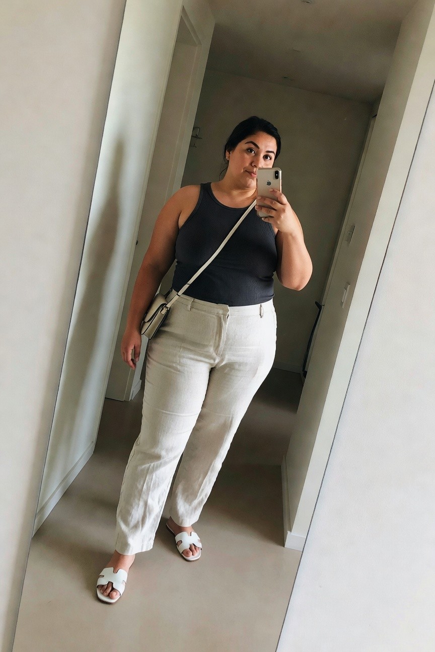 Woman in black tank top and white pants taking mirror selfie with phone.