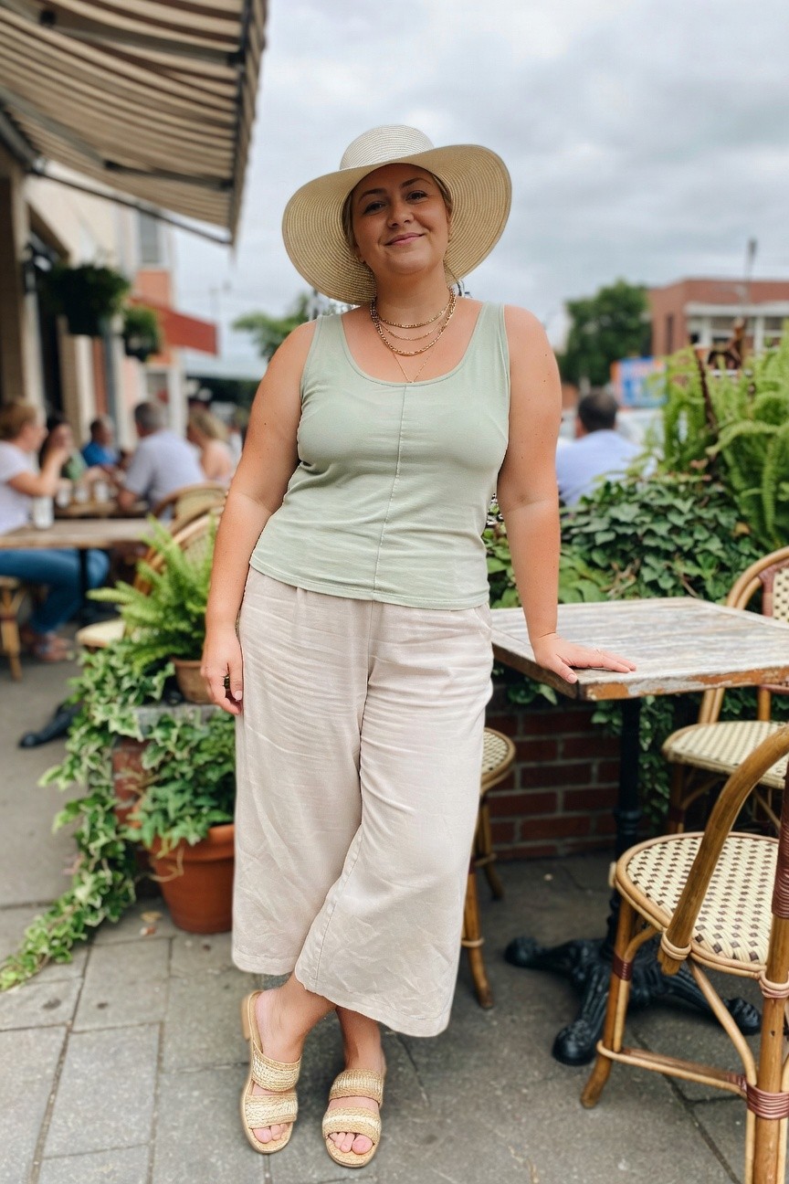 Plus size woman in a sage green scoop neck tank top with a center seam and beige wide leg cropped pants, layered gold necklaces, woven slide sandals and a wide brim straw hat, standing beside an outdoor table.