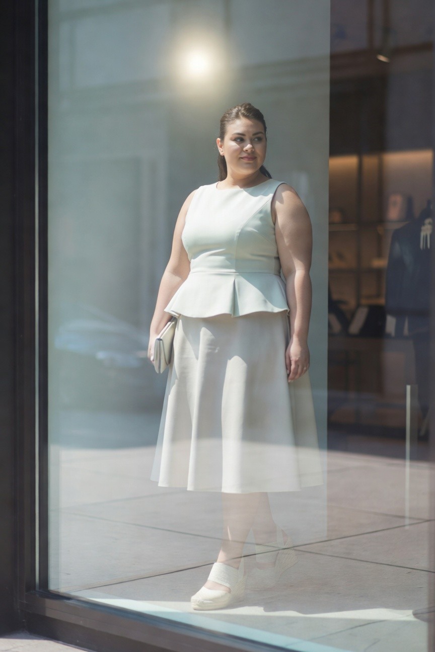 Woman in light green peplum top and flared skirt standing by glass window.