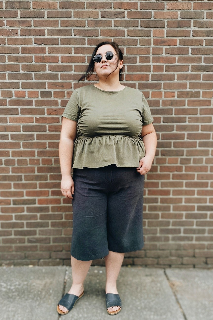 Woman in olive peplum top and dark shorts stands against brick wall.