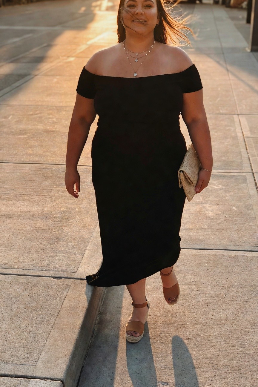 Woman in black off-shoulder dress holding beige clutch on sunny sidewalk.