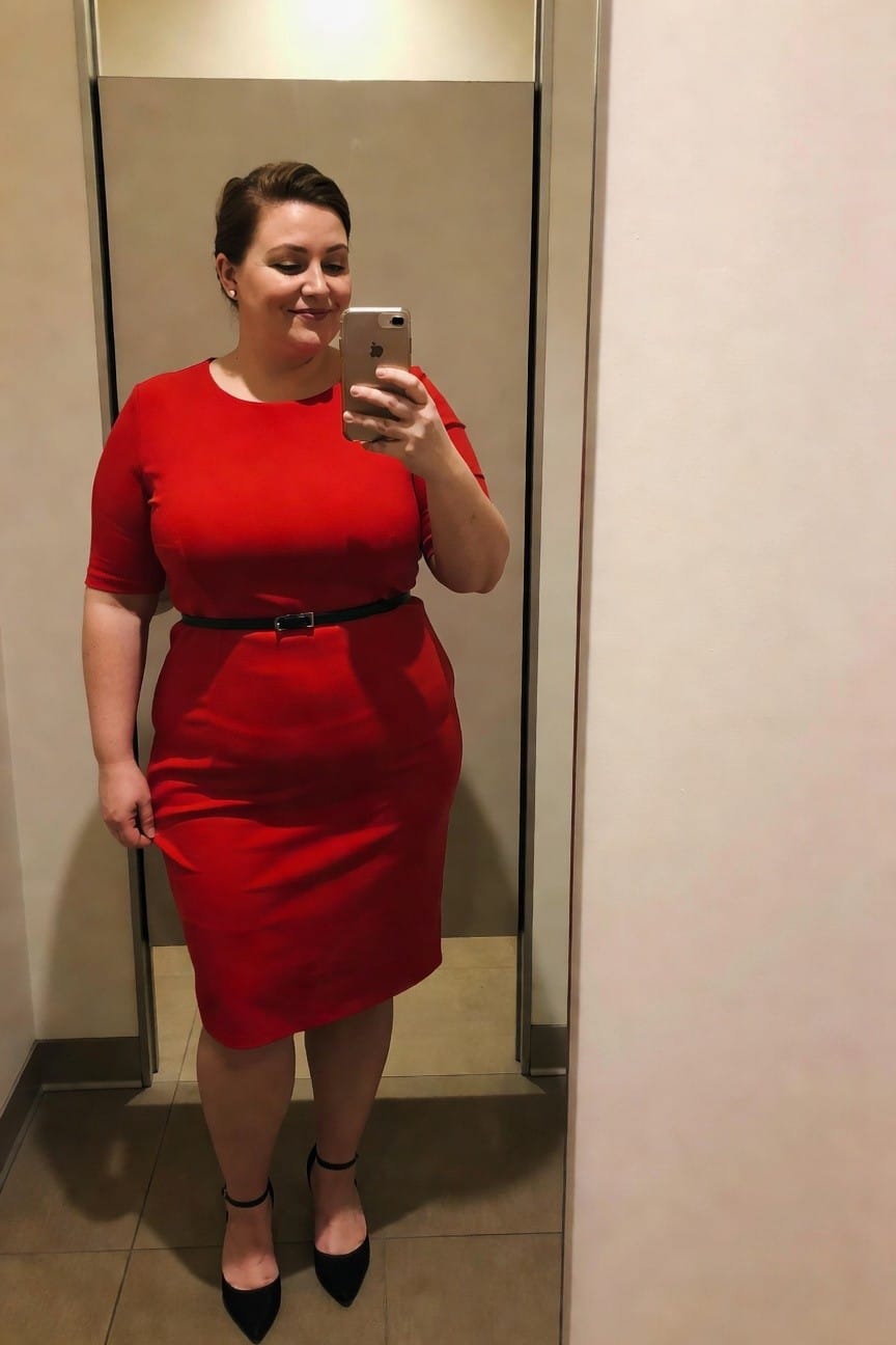 Plus size woman in a fitted red midi dress with short sleeves and black belt taking a mirror selfie while wearing black ankle strap heels.