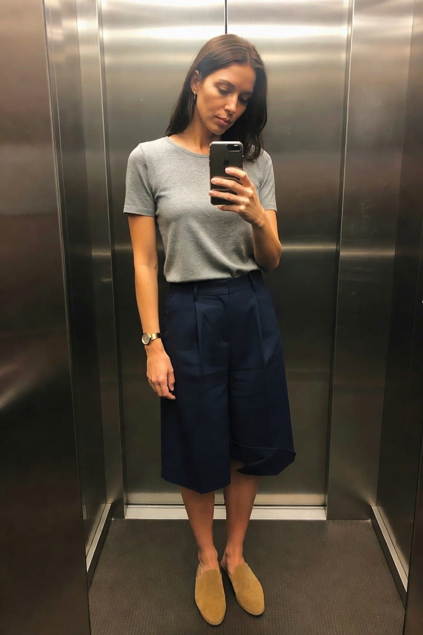 Woman wearing a gray short sleeve tee with navy wide leg culottes and beige suede loafers while holding a phone in an elevator selfie.