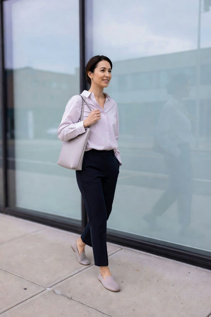 Woman in lavender blouse and navy pants holds beige handbag by glass building.