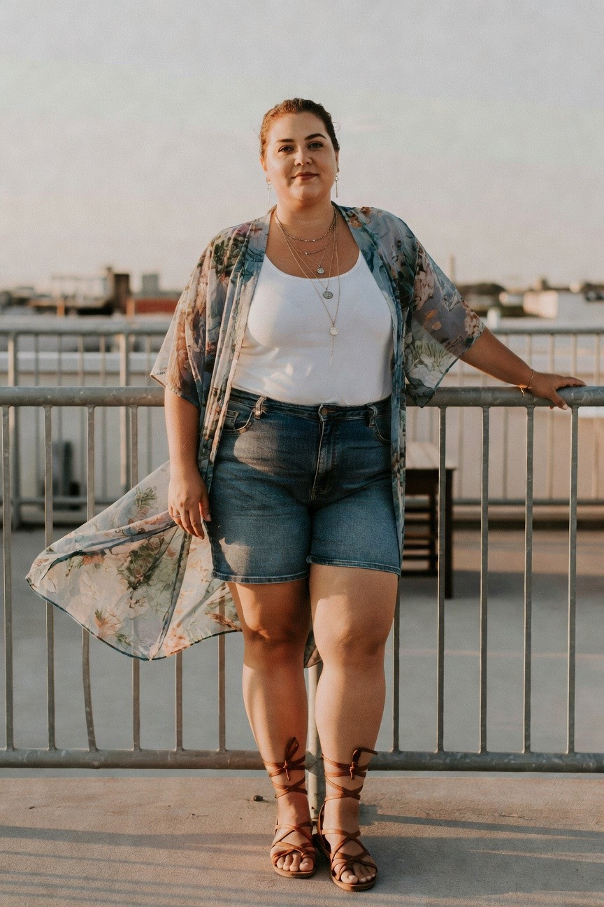 Woman in floral kimono, white tank, and denim shorts standing by railing.