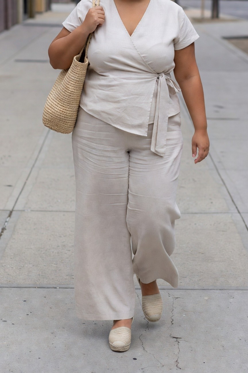 Woman in beige linen wrap top and wide-leg pants carrying woven bag.
