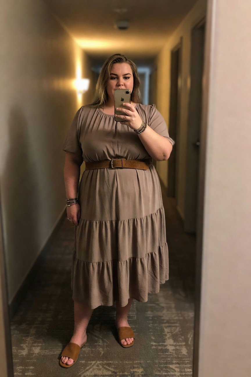 Woman in taupe tiered dress taking a mirror selfie in a hallway.