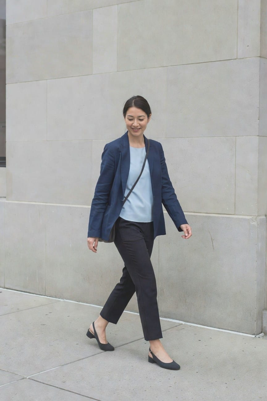 Woman in navy blazer over light blue top with dark pants and black slingback flats walking on pavement