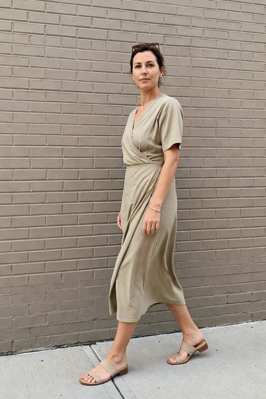 Woman in beige wrap dress and sandals against a gray brick wall.