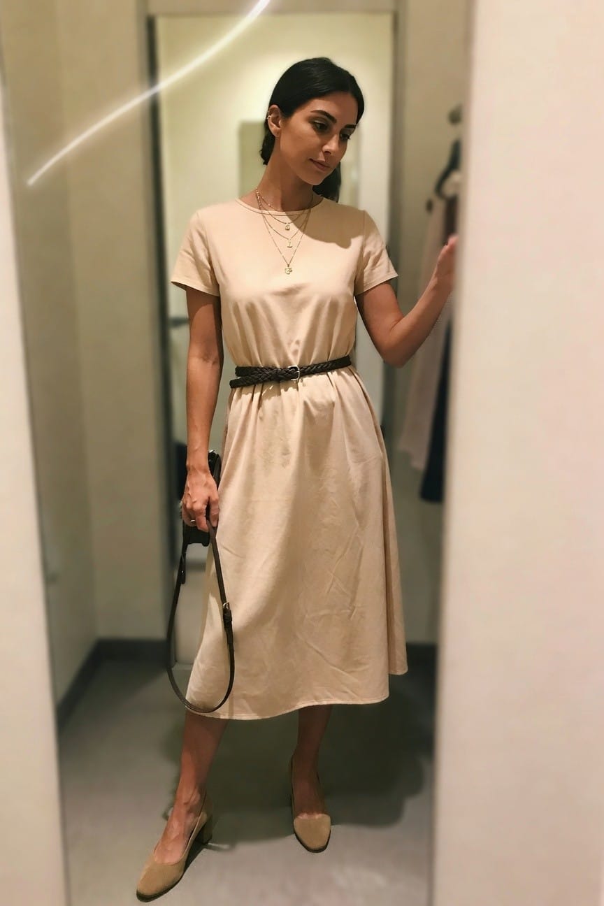 Woman in beige midi dress with braided belt and layered necklaces holding a handbag.