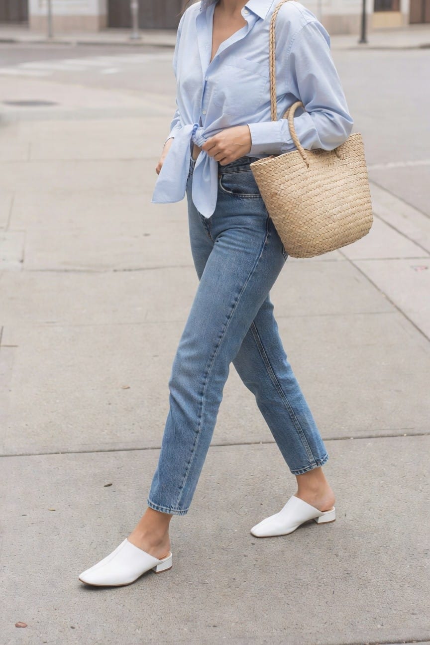 Woman in light blue shirt and jeans with woven straw bag and white mules.