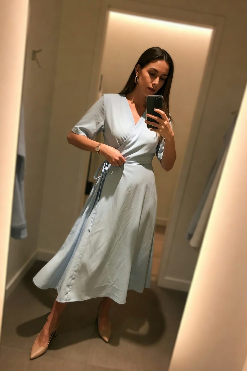 Woman in light blue wrap dress taking mirror selfie with phone.