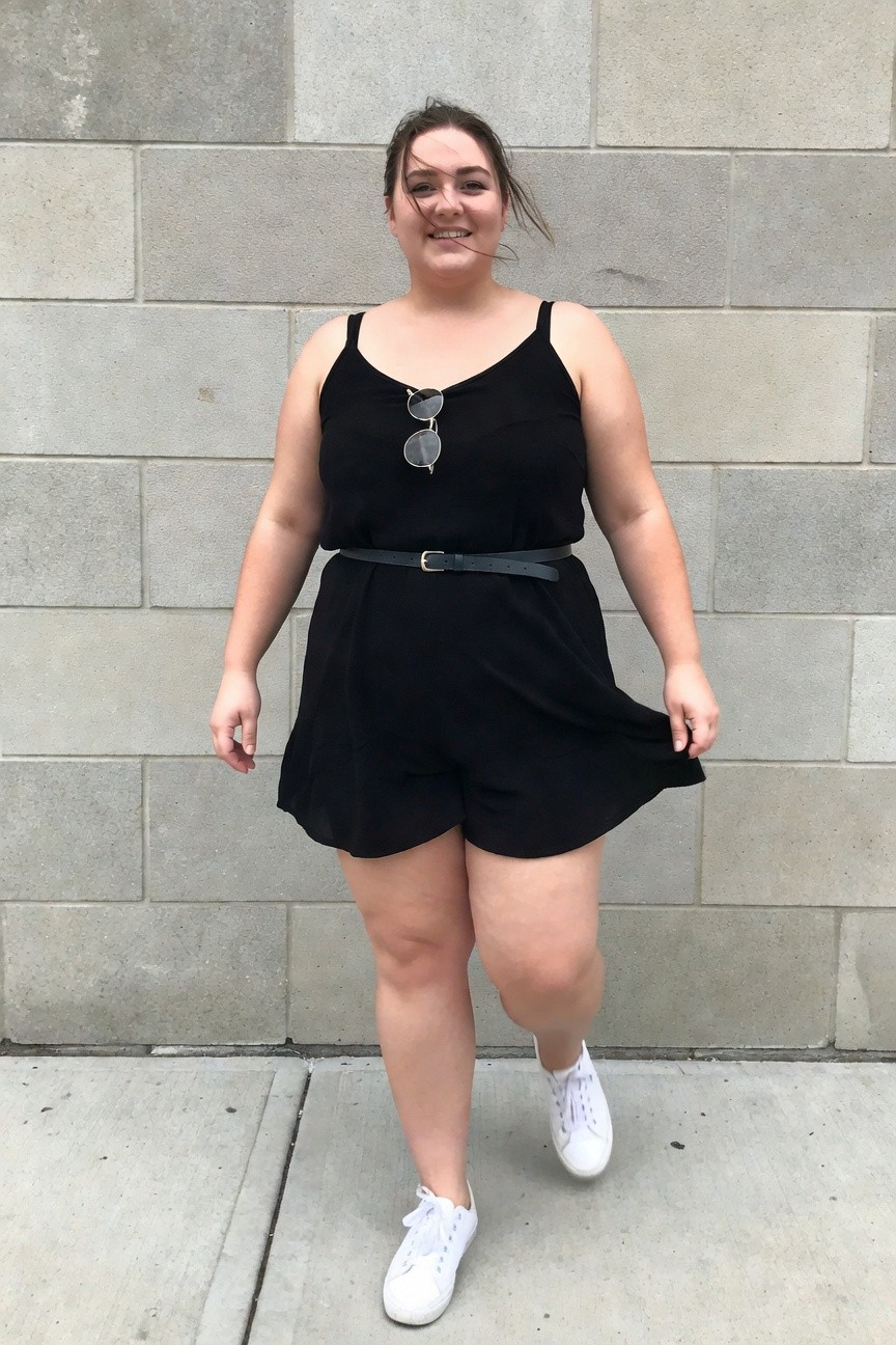 A smiling woman in a black romper with white sneakers and sunglasses on her chest.