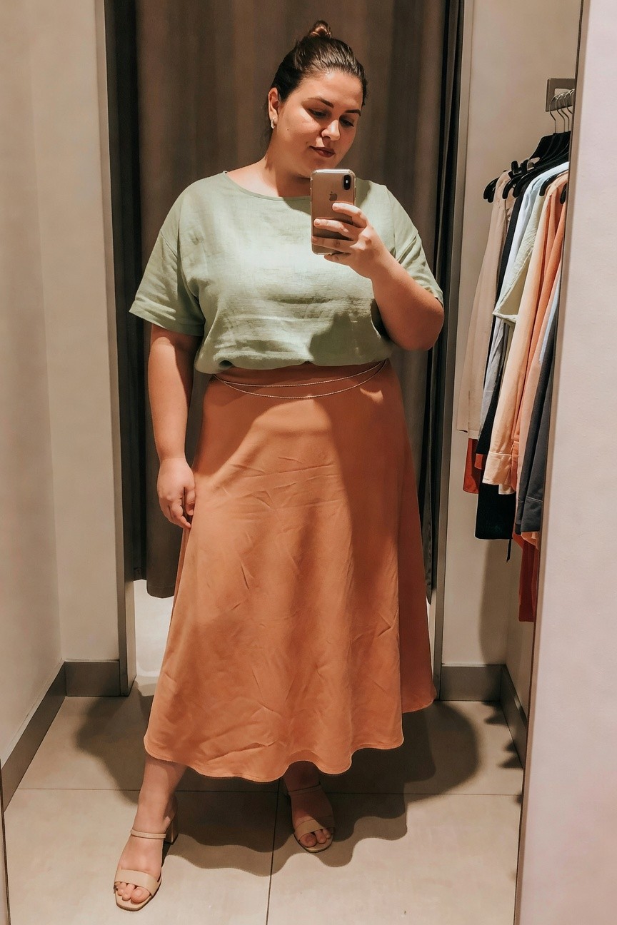 Plus size woman wearing a sage green short sleeve linen top tucked into a terracotta orange midi skirt with a thin silver chain belt and beige block heel sandals while taking a mirror selfie in a fitting room.