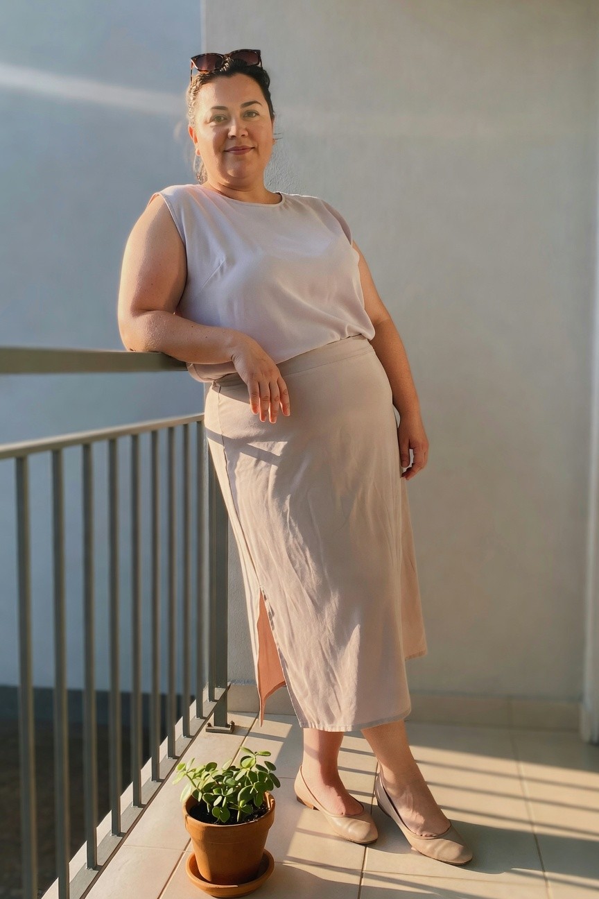 Plus size woman wearing a light beige sleeveless top tucked into a matching midi skirt with side slit and nude flats, sunglasses resting on her head.