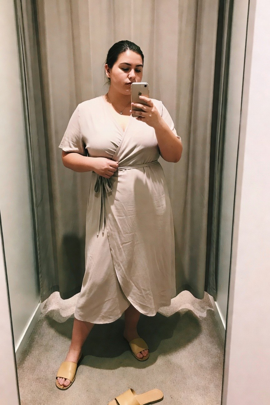 Woman in beige wrap dress taking a mirror selfie with phone.