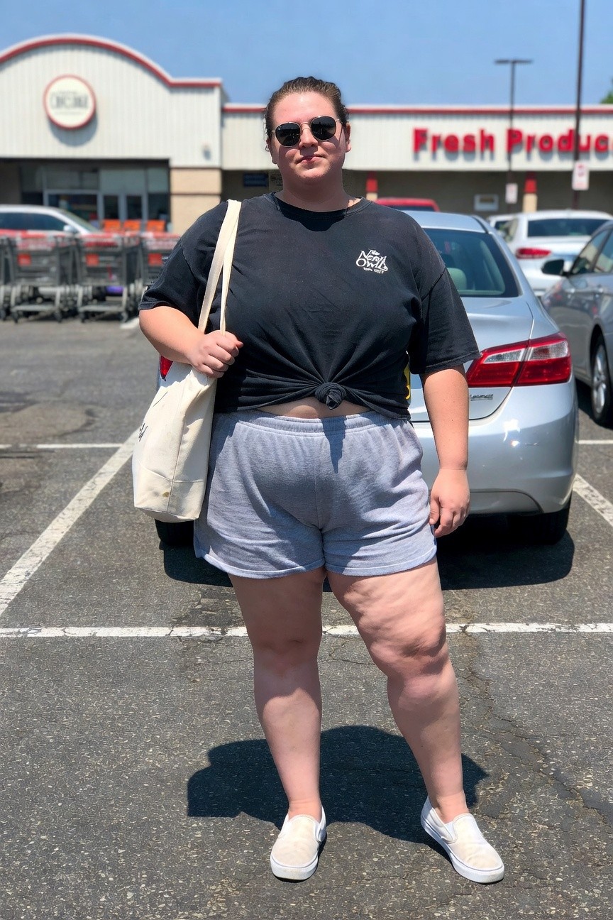 Woman in black t-shirt and gray shorts standing in sunny parking lot.