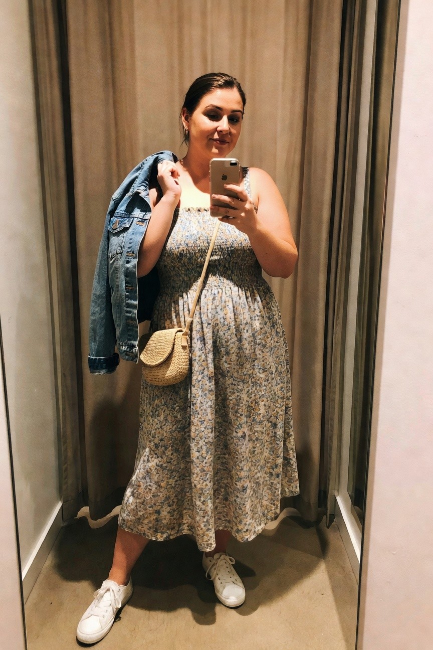 Woman in floral dress taking mirror selfie with denim jacket and woven bag.