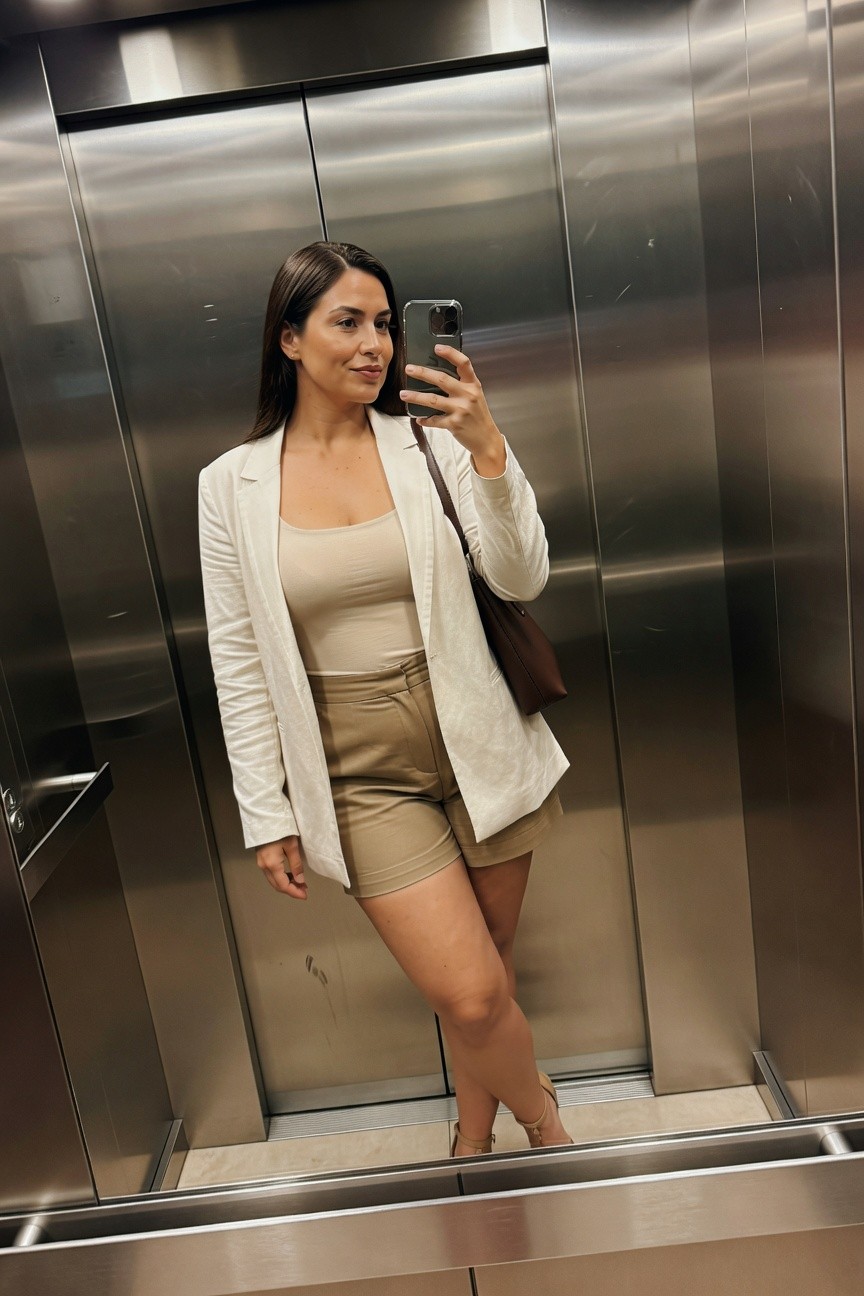 Woman in beige outfit taking mirror selfie inside elevator.