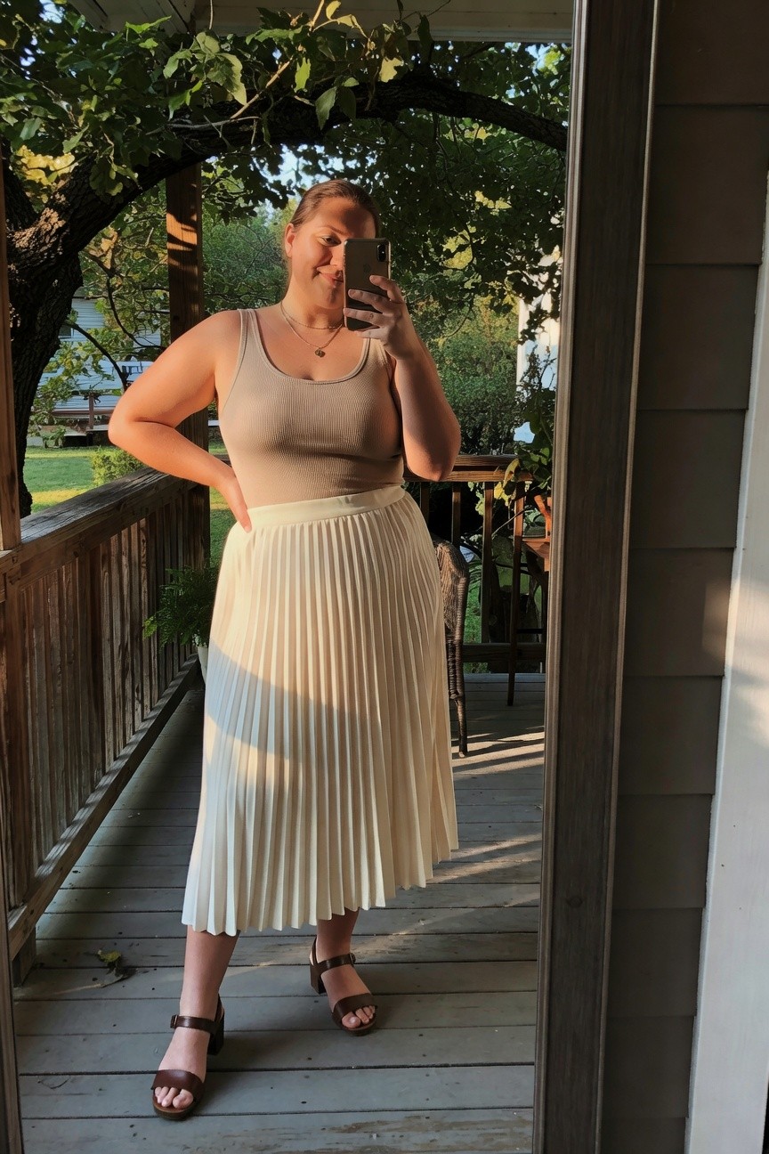 Woman in beige tank top and pleated cream skirt taking a mirror selfie.