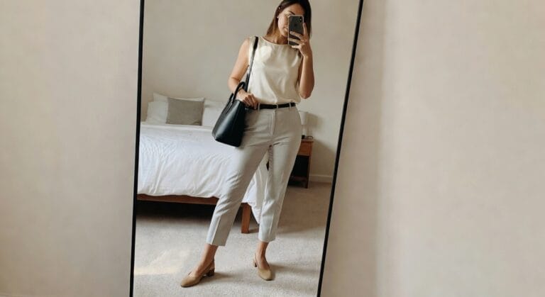 A woman poses in front of a mirror, wearing a cream sleeveless top and light gray tailored pants, complemented by a black handbag and beige flats.