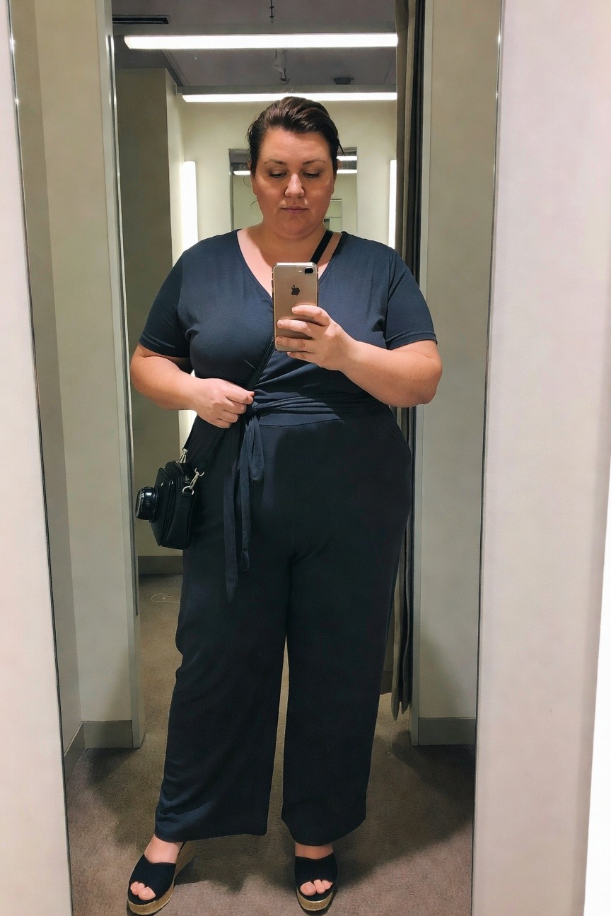 Plus size woman wearing a navy short sleeve jumpsuit with tie waist detail and wide legs, small black crossbody bag, black wedge sandals, taking a mirror selfie with phone.