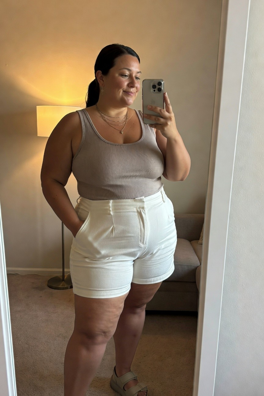 Woman in beige tank top and white shorts taking a mirror selfie.