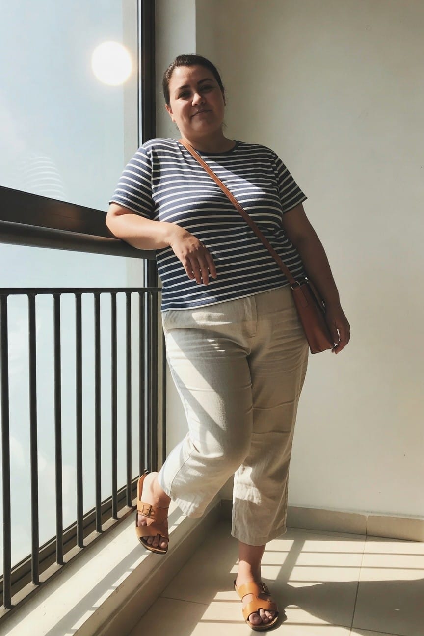 Woman in striped shirt and beige pants leaning by sunny window.