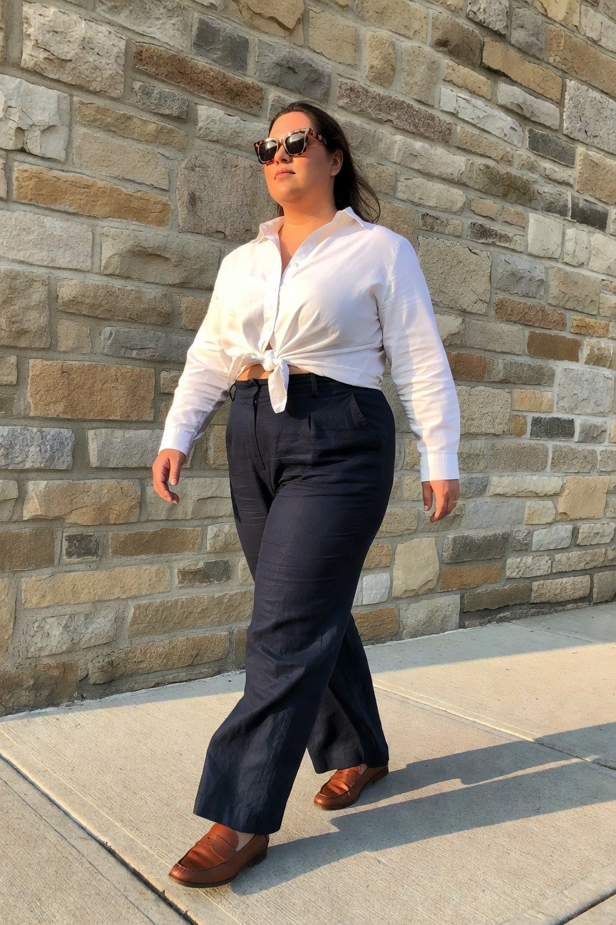 Woman in white tied shirt and navy pants posing before stone wall.