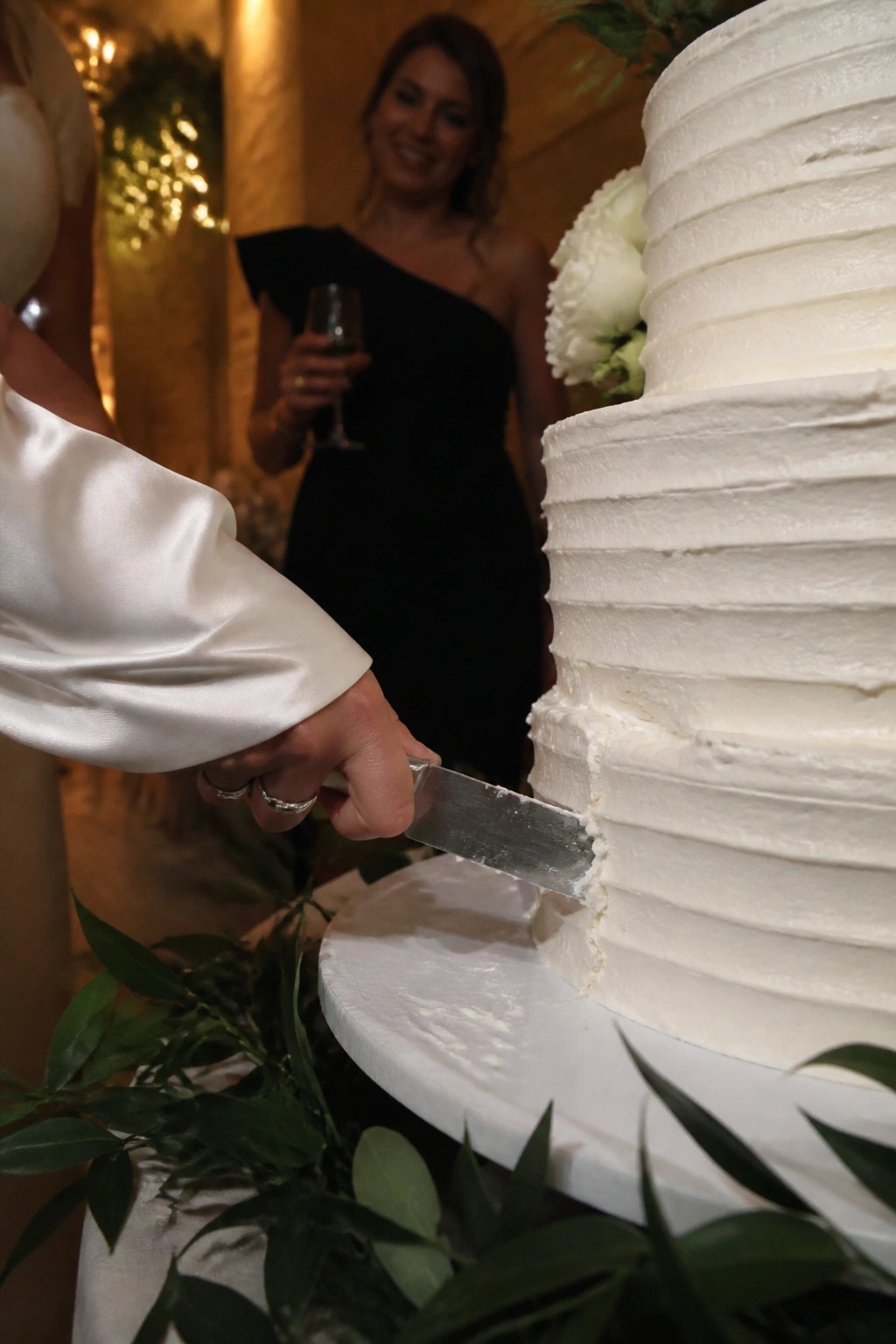 Close up of hands in white satin cutting a multi tiered white wedding cake with a smiling guest in black holding champagne in the background.