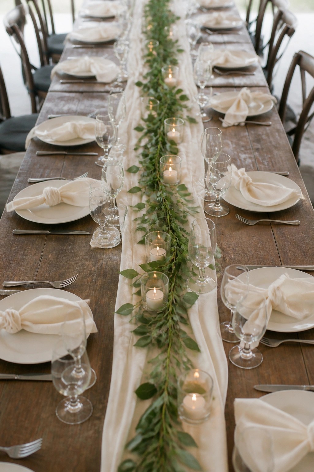 A long wooden wedding table set with white plates, knotted napkins, wine glasses, and a central runner of green foliage interspersed with lit candles in glass votives. The scene features a neutral color palette with wooden chairs and soft lighting.