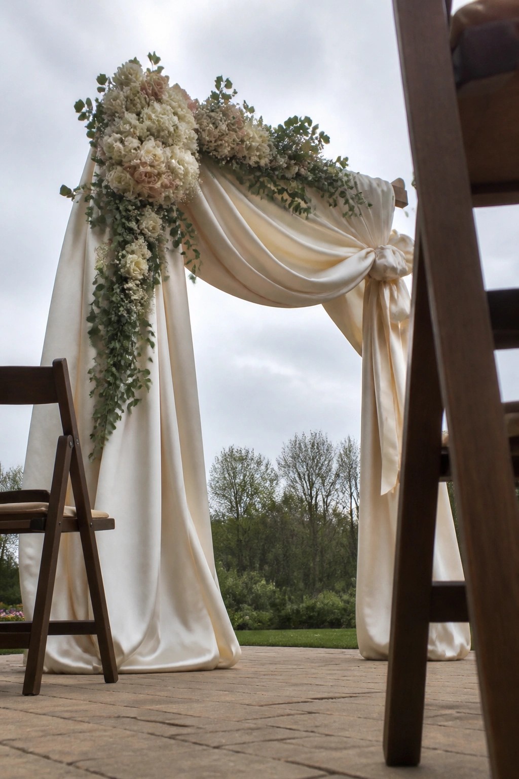 Wedding arch draped in white satin with white and blush floral arrangements and greenery on a brick patio beside wooden chairs.