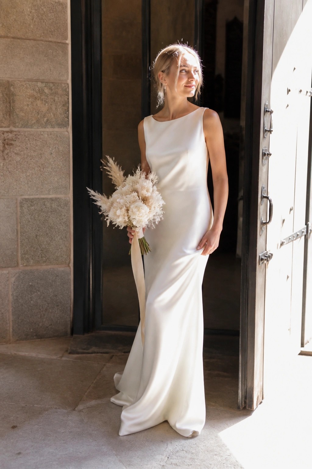 Bride in form fitting white satin gown holds neutral floral bouquet while standing in open wooden doorway of stone building with sunlight illuminating the scene