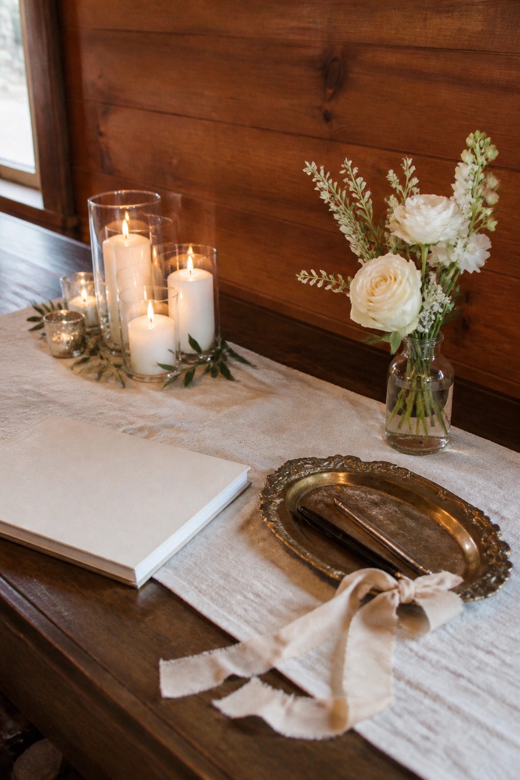 A wooden table with white linen runner displays glowing pillar candles in glass holders, a vase of white roses, a gold pen tray with satin ribbon, and an open guest book against a wood wall.