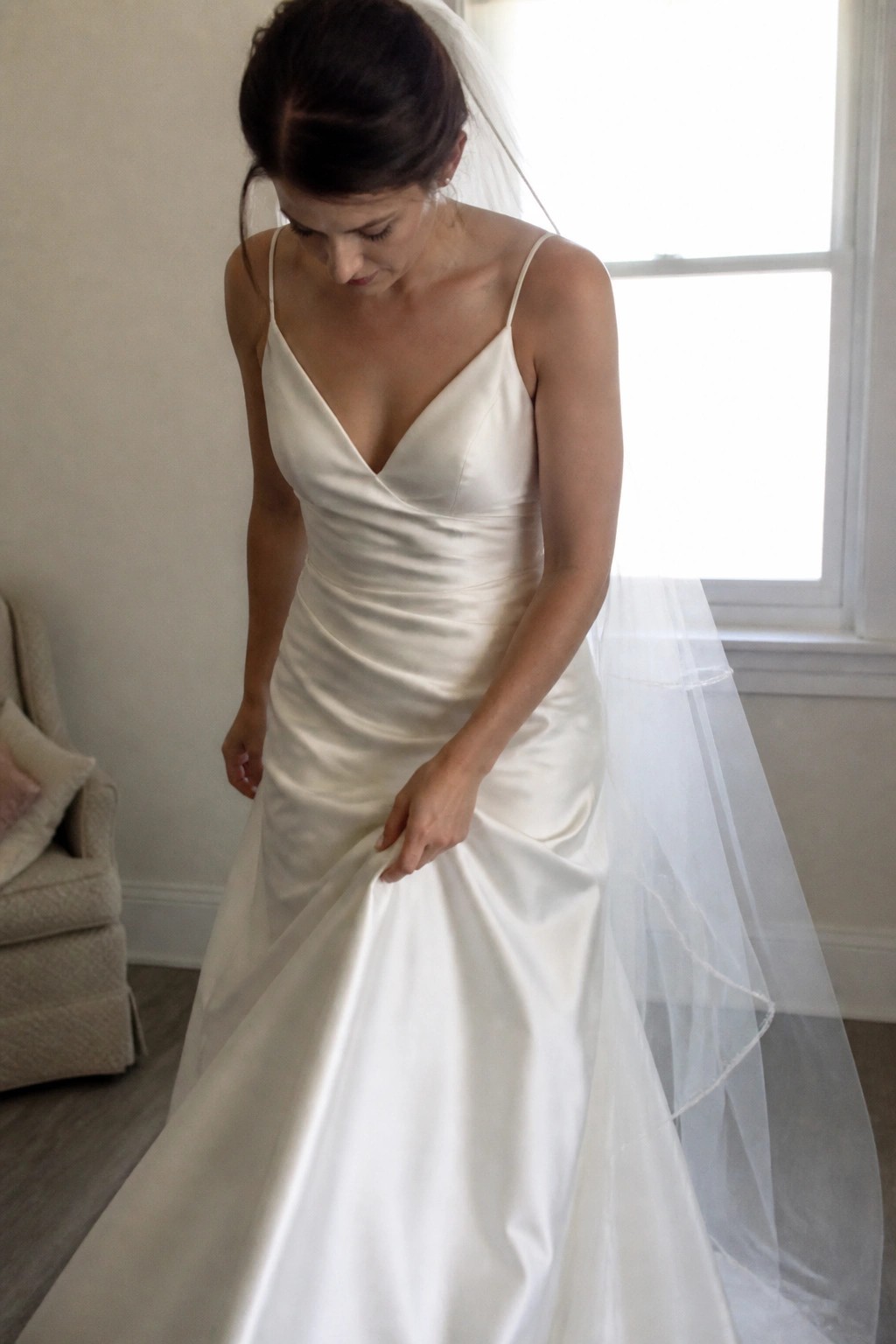 Bride in ivory satin wedding dress adjusts her skirt while wearing a sheer veil in a sunlit neutral room.