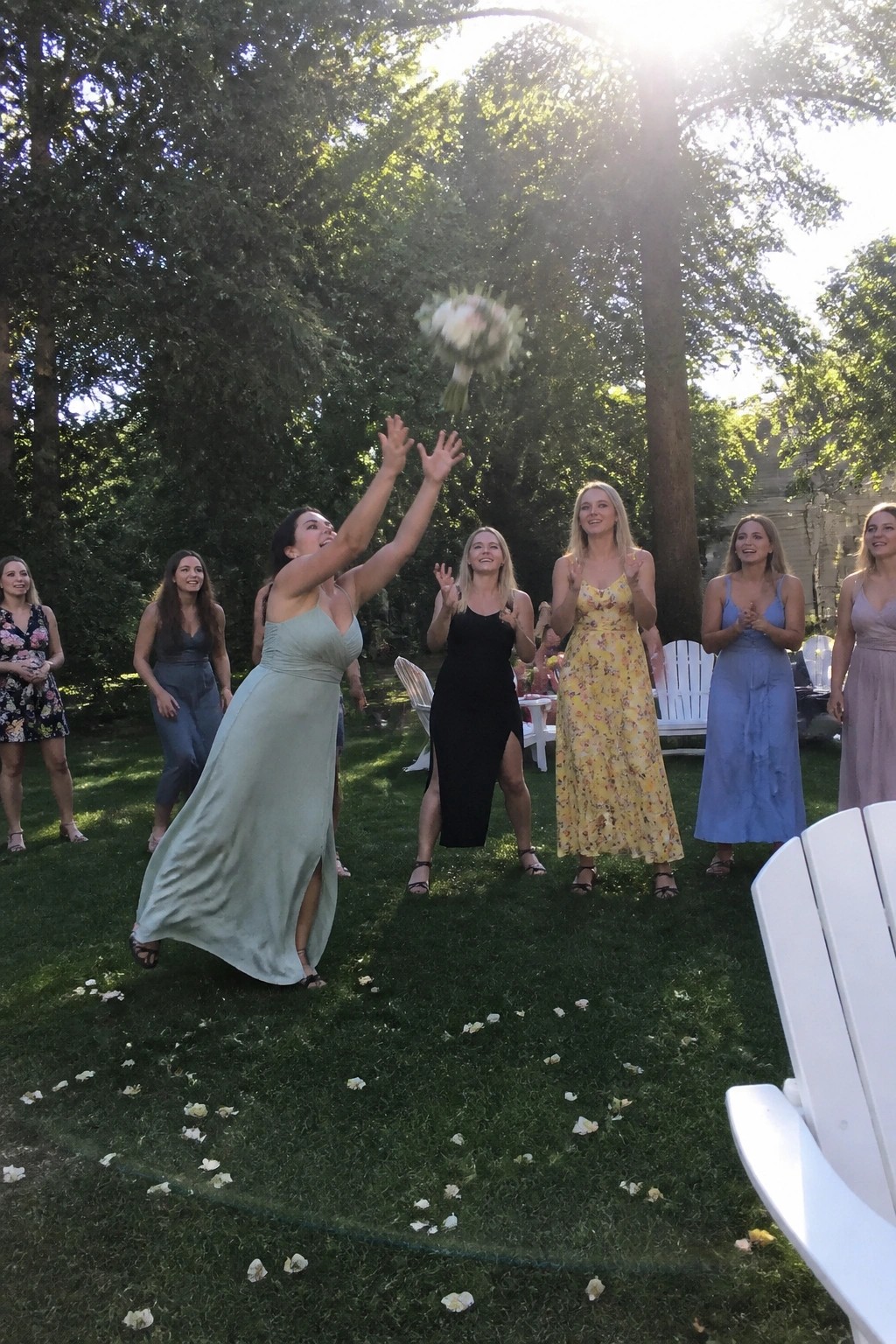 Bridesmaids leap and cheer during an outdoor bouquet toss on a sunny lawn with trees and white chairs in the background.