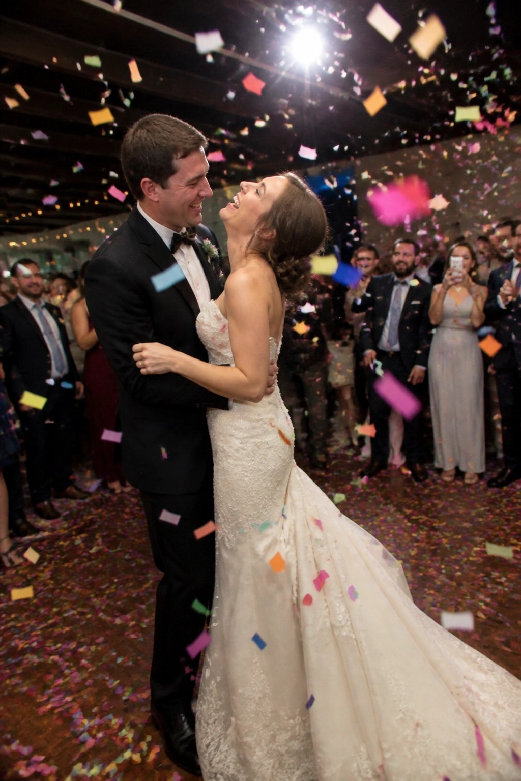 Bride and groom embrace amid falling colorful confetti at their wedding reception with smiling guests in the background.