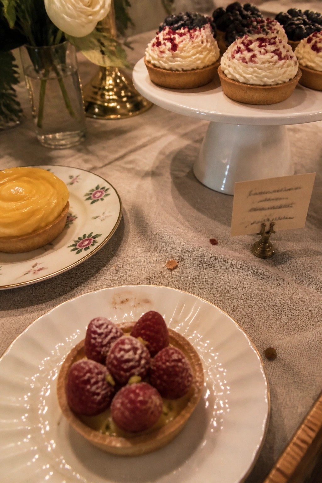 Close up of elegant wedding dessert tarts including strawberry, lemon, and berry varieties arranged on vintage plates and a cake stand with floral accents on a linen table.