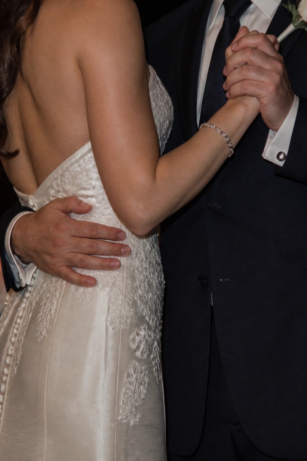 Close up of bride in white lace gown and groom in dark suit holding hands with his hand on her waist from behind