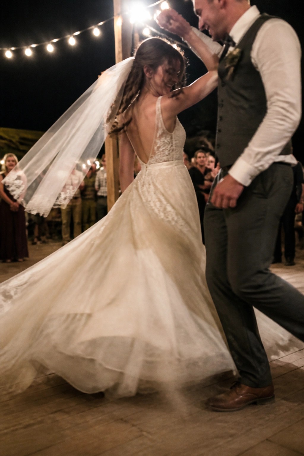 Bride in lace gown with flowing veil twirls joyfully as groom spins her under string lights at outdoor night reception on wooden floor with guests watching in background.