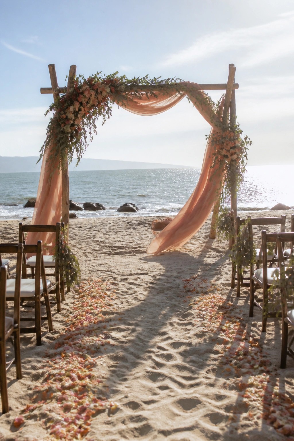Wooden beach wedding arch draped in flowing peach fabric with peach floral accents and greenery overlooking the ocean with petal lined aisle and wooden guest chairs